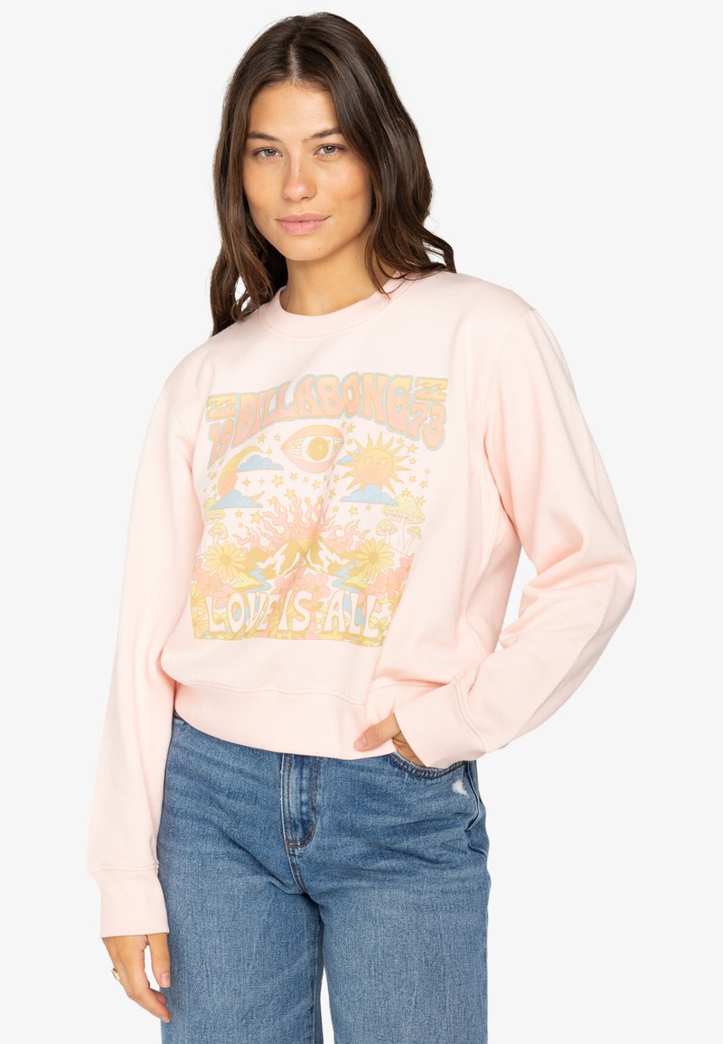 Rosa Sweatshirt mit einem bunten Grafikdruck, der Sonne, Auge und florale Motive zeigt. Rippbündchen und Saum. Kombiniert mit hellen Jeans.