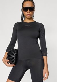 Černý dlouhý cyklistický dres s kulatým výstřihem a logem; doplněný černými cyklistickými kraťasy s polstrovaným sedlem. Model drží černou helmu.