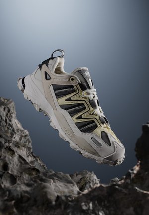 Valkoinen ja musta Adidas-tennari karkealla pohjalla, kellumassa kallioisen pinnan yläpuolella harmaan liukuvärisen taustan edessä.