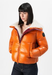 Orange puffer jacket with a shiny exterior, fleece-lined hood, purple zipper detailing, and a round black patch on the left sleeve.
