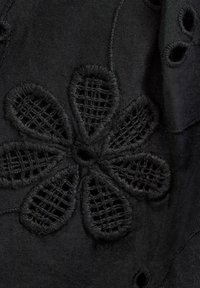 Svart textil med blommig broderi och utskurna detaljer, som visar texturerade blad och ett centralt hål, vilket skapar en lagerdesign.