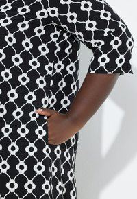 Czarna sukienka z białym kwiatowym wzorem, z kieszenią po lewej stronie i rękawami do połowy ramienia. Gładkie wykończenie tkaniny.