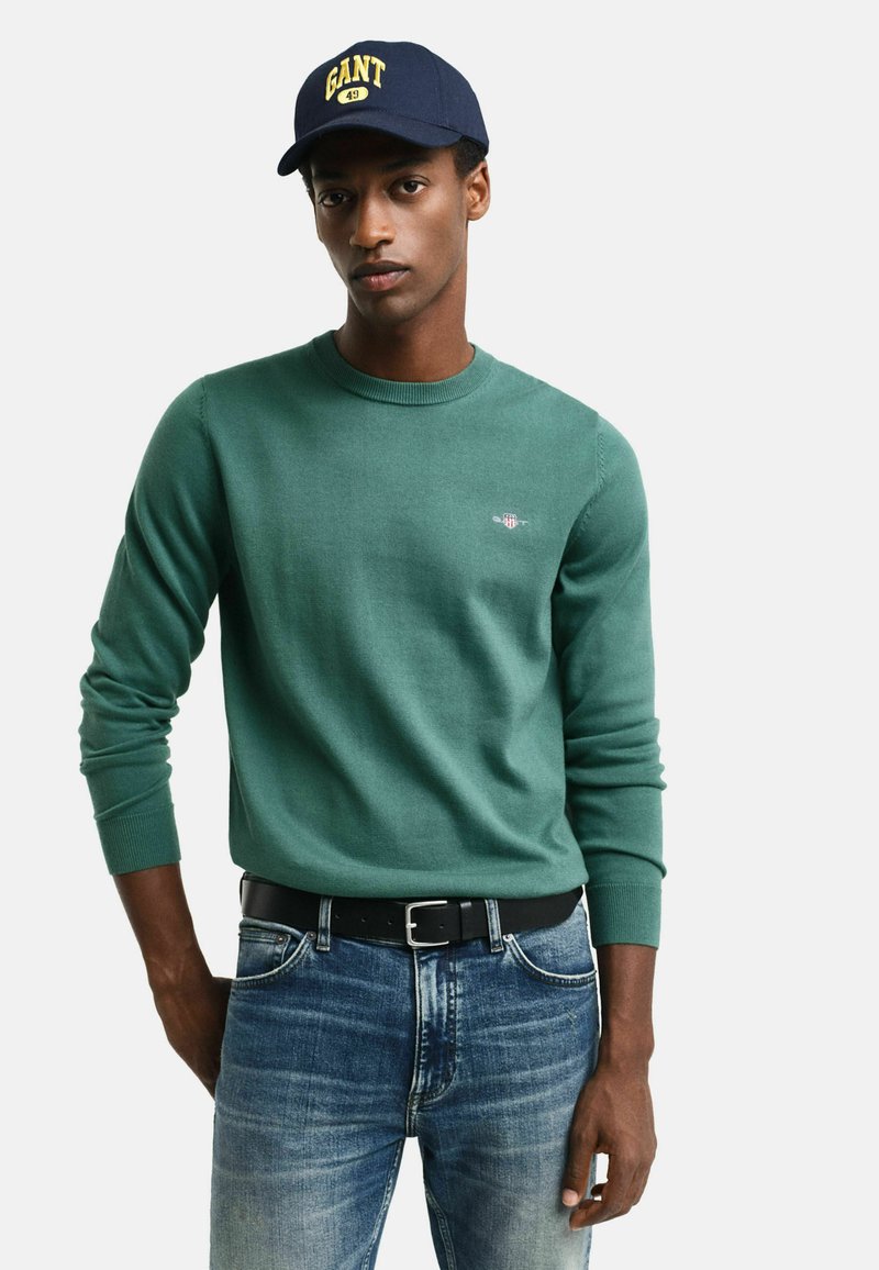Grüner Pullover mit Rundhalsausschnitt, langen Ärmeln und kleinem Logo auf der Brust, kombiniert mit blauen Jeans und einem schwarzen Gürtel.