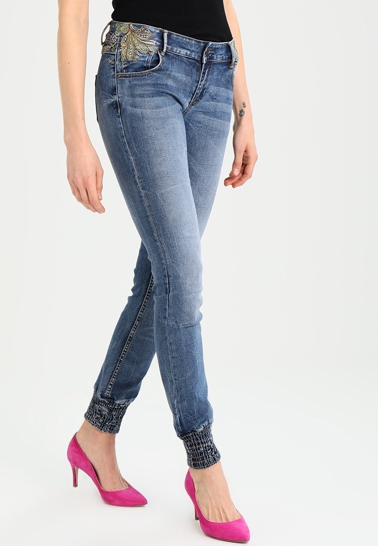 Femme portant un jean bleu slim avec un détail brodé à la taille et des chevilles retroussées, associées à des talons hauts rose vif.