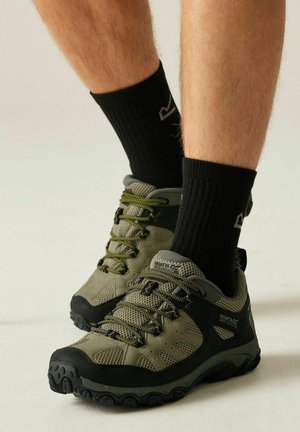 Męskie stopy w oliwkowo-czarnych butach do wędrówek i czarnych skarpetkach na jasnym tle.