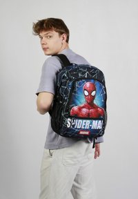 Un sac à dos noir avec un grand motif de Spider-Man, un motif de toile bleu, des zips doubles et des bretelles ajustables. Le matériau semble durable.
