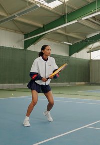Witte sportjasje met marine accenten, geplooide marine rok en witte sneakers; houdt een tennisracket binnen op een blauwe vloer.