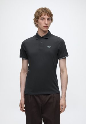 Jeune homme aux cheveux blonds bouclés, portant un polo noir Barbour et un pantalon foncé, debout devant un fond blanc uni.