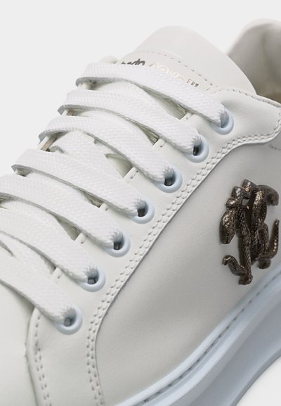 Κοντινό πλάνο ενός λευκού αθλητικού παπουτσιού με λευκά κορδόνια, μεταλλικές οπές και μεταλλικό λογότυπο φιδιού στο πλάι κοντά στη σόλα.