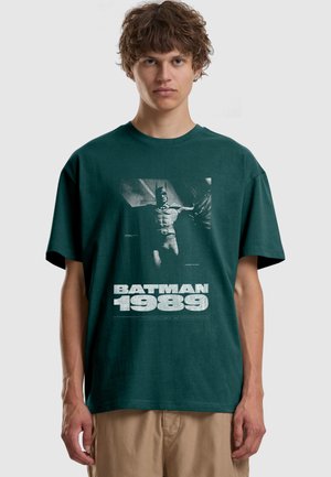 Jonge man met krullend bruin haar die een donkergroene Batman 1989 grafische t-shirt en beige broek draagt tegen een effen achtergrond.
