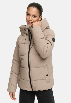 Vrouw in een beige gewatteerde jas met zwarte rits en capuchon, die de kraag van de jas vasthoudt, staand tegen een effen achtergrond.