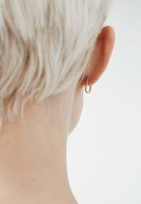 Pendiente de aro dorado exhibido sobre un fondo de cabello corto y claro. El diseño presenta una forma lisa y minimalista.