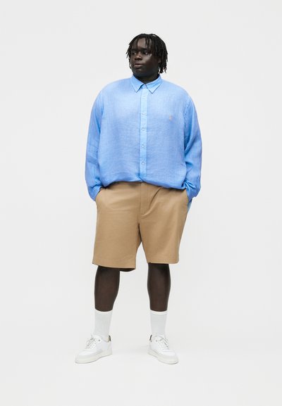 Vyras tamsia oda ir dredais, vilkintis laisvą šviesiai mėlyną marškinių tipo palaidinę, smėlio spalvos šortus, baltas kojines ir baltus sportinius batelius, stovintis rankomis kišenėse.