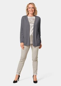 Grijze gebreide cardigan met een open voorkant en twee zakken, gedragen over een lichte top. Gecombineerd met beige broek en zwarte hoge hakken.