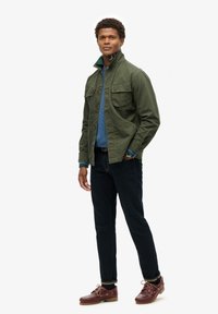 Donkergroene, lichte jas met twee borstzakken over een blauwe trui, gecombineerd met donkere spijkerbroeken en bruin leren schoenen met veters.