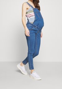 Grå denimoverall i blått för gravida, med justerbara axelremmar, framfickor och en avslappnad passform, kombinerad med vita sneakers.