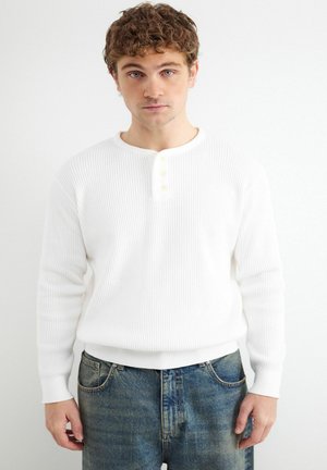 Młody mężczyzna z kręconymi włosami, ubrany w białą prążkowaną koszulkę henley z długim rękawem i niebieskie dżinsy na białym tle.