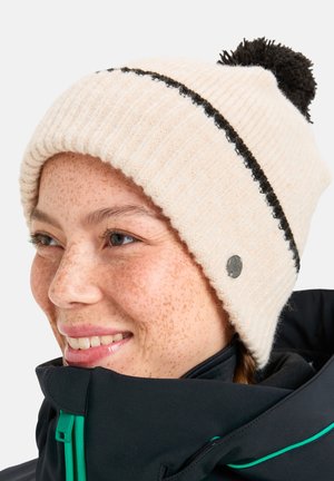 Jeune femme aux freckles souriante, portant un bonnet en tricot crème avec un pompon noir et une veste d'hiver noire avec des détails de fermeture éclair verts.