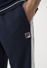 Navyfarbene Sport-Jogginghosen mit seitlichen weißen Streifen, die auf der Gesäßtasche ein markantes rechteckiges Logoschild in Rot, Weiß und Schwarz aufweisen.
