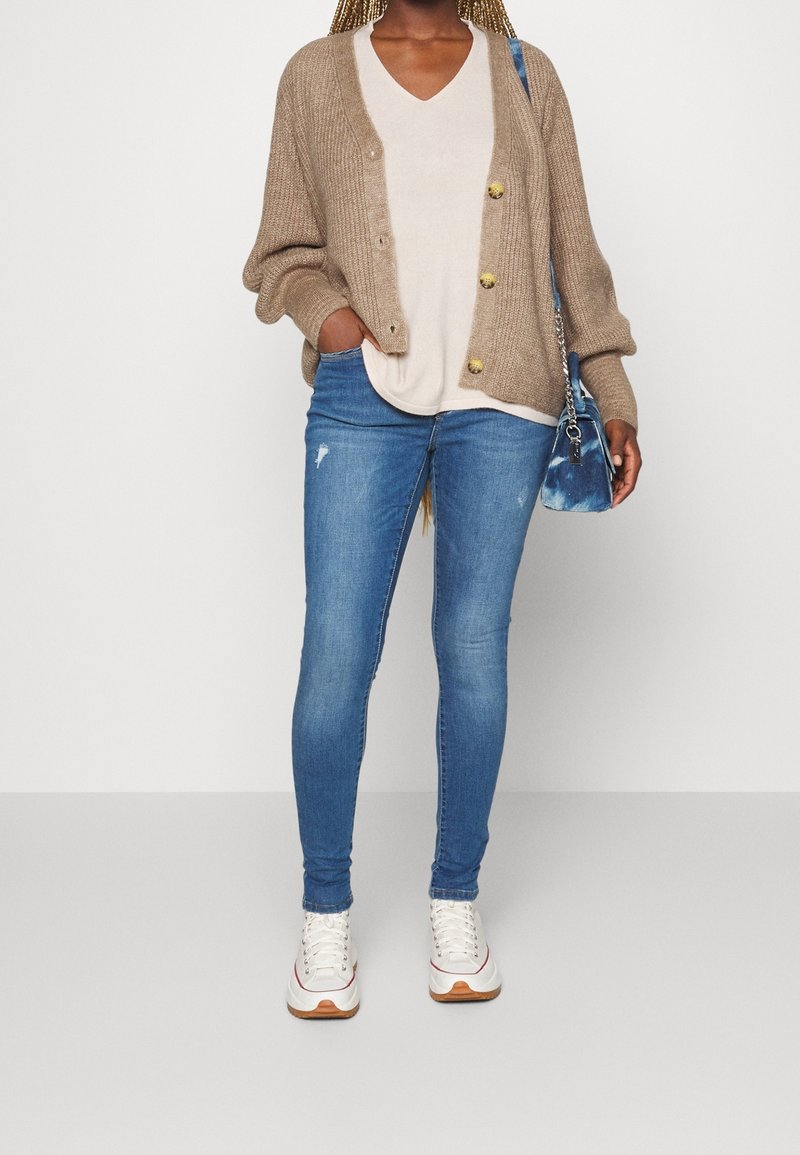 Personne portant un cardigan beige sur une chemise blanche, un jean skinny bleu avec légère usure, des baskets blanches, et tenant un sac à main bleu sur le côté.