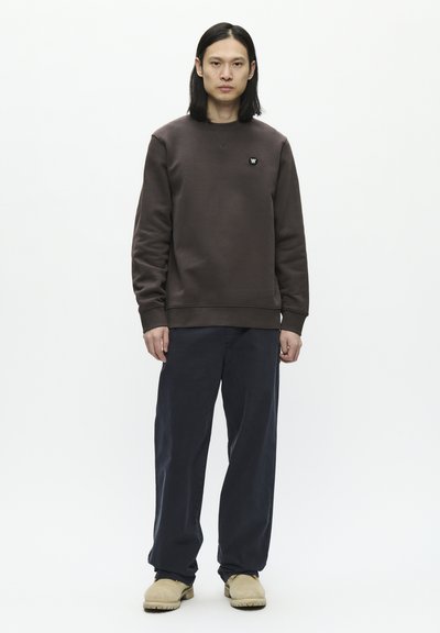 Sweatshirt marron avec des poignets et un col côtelés, associé à un pantalon bleu marine ample et des chaussures à enfiler beiges. Pas de motifs visibles.