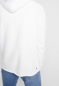 Felpa bianca con finitura strutturata, dotata di cappuccio con coulisse, maniche raglan e orlo arrotondato. Include un bottone decorativo in metallo.