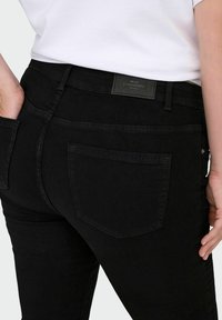 Fekete farmernadrág sima textúrával, hátsó zsebbel és márkázott felirattal a deréknál. Közelkép a hátsó nézetről.