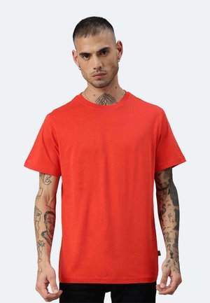 Mann mit kurzen Haaren und Tätowierungen an Hals und Armen, trägt ein schlichtes leuchtend oranges kurzärmeliges T-Shirt und steht vor einem hellen Hintergrund.