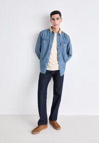 Light blue denim shirt with two chest pockets, cream crew neck shirt underneath, dark blue jeans, and brown sneakers. Standing pose against white background.