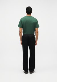 Άνδρας που στέκεται γυρισμένος από πίσω, φορώντας πράσινο κοντομάνικο πουκάμισο, μαύρο παντελόνι και σκούρα παπούτσια, μπροστά σε απλό λευκό φόντο.