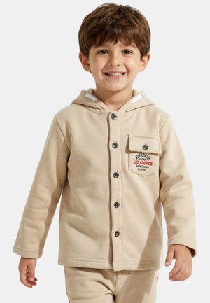 Jeune garçon souriant, portant une veste à capuche beige à boutons avec une poche poitrine et un pantalon assorti, sur un fond uni.