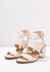 Beige korkokengät, joissa on sileä nahkainen yläosa, leveät strapsit ja teksturoitu tausta. Varustettu paksulla metallikorkolla, jossa on neliön muoto.