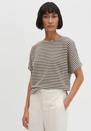 Donna con capelli corti neri che indossa una maglia larga a maniche corte a righe nere e beige e pantaloni bianchi a vita alta, posando su sfondo bianco.