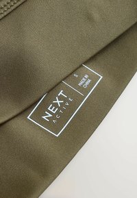 Tissu de vêtements de sport vert olive avec une texture lisse. Étiquette blanche portant l'inscription « NEXT ACTIVE », taille « S » et « FABRIQUÉ EN CHINE » imprimée en dessous.