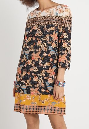 Femme portant une robe fleurie mi-longue avec des sections noir, blanc et jaune moutarde, ainsi qu'un bracelet coloré à motifs à son poignet.