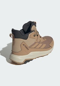 Beige Wanderschuh mit einem strukturierten Obermaterial, verstärktem Absatz, schwarzen Akzenten und einer dicken Sohle. Verfügt über einen Ziehgriff für einfaches Anziehen.