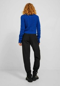 Afgeknipte blauwe gebreide trui met gestructureerd patroon, gecombineerd met loszittende zwarte broek en zwarte chunky schoenen, gezien van achteren.