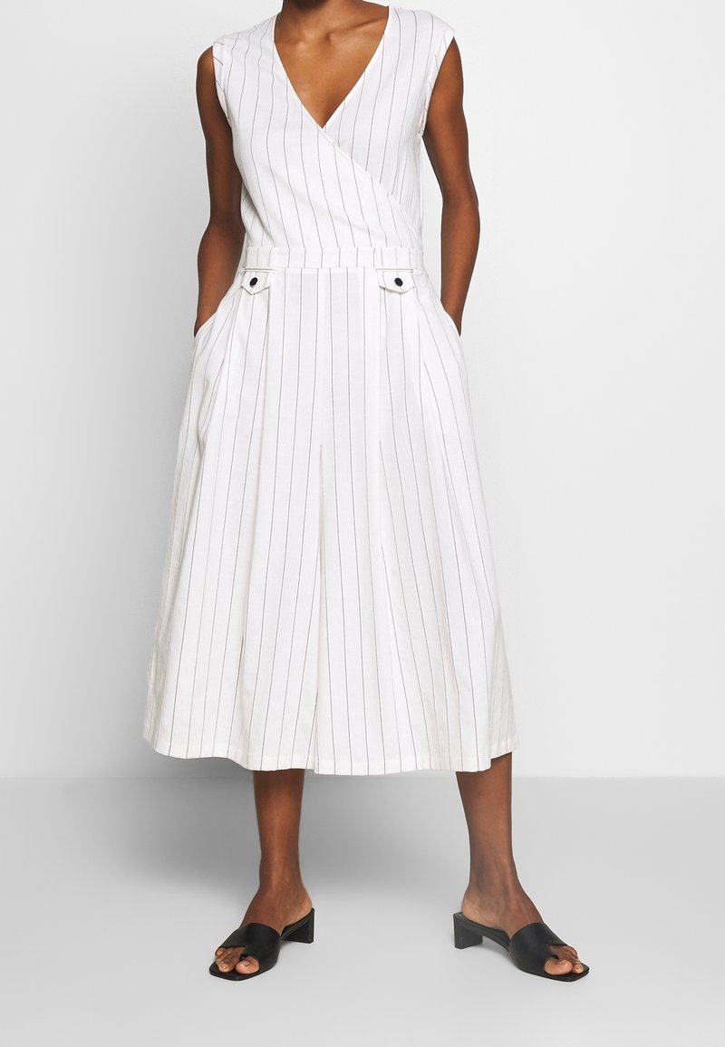 Robe midi blanche sans manches avec fines rayures verticales noires, encolure en V, poches avant, portée avec des sandales noires à talons blocs et bouts ouverts.