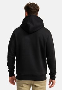 Mężczyzna w czarnej bluzie z kapturem, kaptur opuszczony, widziany od tyłu, stoi na jasnym, jednolitym tle.