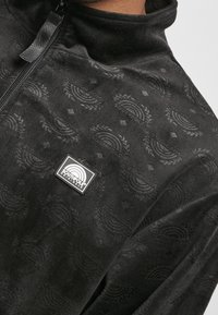 Svart fleece-pullover med hög krage, med ett strukturerat mönster av bågar och diamanter. Inkluderar ett logomärke på bröstet.