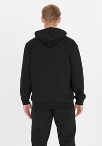 Mies, joka katsoo poispäin, lyhyet vaaleat hiukset, päällään musta huppari ja matching mustat housut valkoista taustaa vasten.