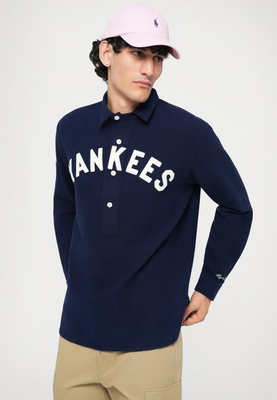 Polo bleumarin cu mânecă lungă, cu text alb "YANKEES", guler cu nasturi și accente brodate. Purtați cu o șapcă roz și pantaloni bej.