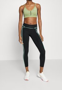 Jasnozielony sportowy biustonosz z regulowanymi ramiączkami, w zestawie z czarnymi legginsami z zielonymi paskami po bokach i logo na pasie, noszonymi z białymi sneakersami.