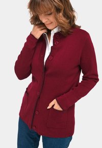Roter Cardigan mit Knopfverschluss, gerippten Bündchen und zwei Vorderstaschen. Akzentnähte entlang des Kragens und der Taschen verleihen Details. Weiches Material.
