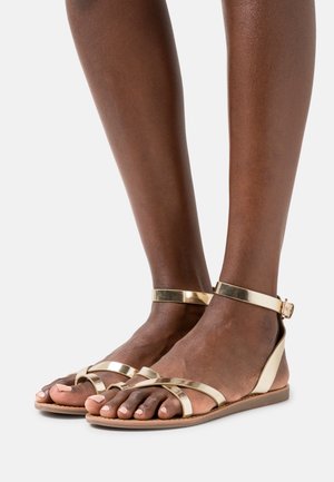 Sandales métalliques dorées à lanières avec boucle au niveau de la cheville, semelle plate et design ouvert au bout. Comprend plusieurs lanières croisées sur le pied.