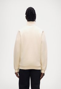 Sweat-shirt beige avec col montant et poignets côtelés, présentant une coupe décontractée et une texture lisse. Porté avec un pantalon sombre.