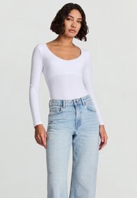 Giovane donna con capelli corti e ricci che indossa una maglia bianca a maniche lunghe con scollo a barca e jeans azzurri a vita alta, davanti a uno sfondo semplice.