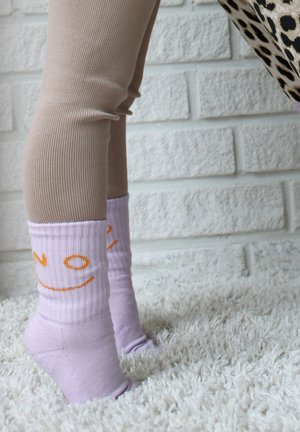 Beine in beige gerippten Leggings und hellvioletten Socken mit orangefarbenen Smiley-Gesichtern stehen auf einem weißen Teppich mit hohem Flor vor einer weißen Ziegelwand.