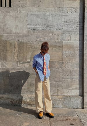 Person iført solbriller, blåstribet skjorte, slips med solsikkemønster, beige bukser og sennepsfarvede sko, står op ad en stenmuret væg med hænderne i lommerne.