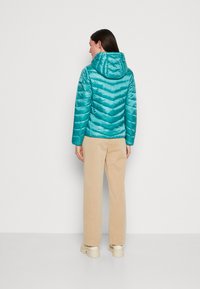 Veste en duvet teal avec un motif en chevrons et une capuche, associée à un pantalon beige clair et des chaussures crème, vue de dos.